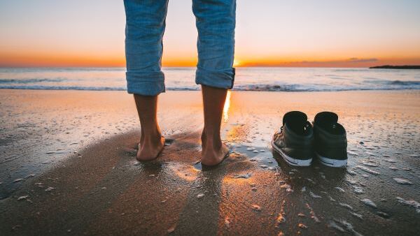 Person steht am Strand in der Abendsonne, die Schuhe ausgezogen und neben sich gestellt - Symbol für einen lockeren Umgang in der schriftlichen Kommunikation