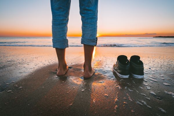 Person steht am Strand in der Abendsonne, die Schuhe ausgezogen und neben sich gestellt - Symbol für einen lockeren Umgang in der schriftlichen Kommunikation